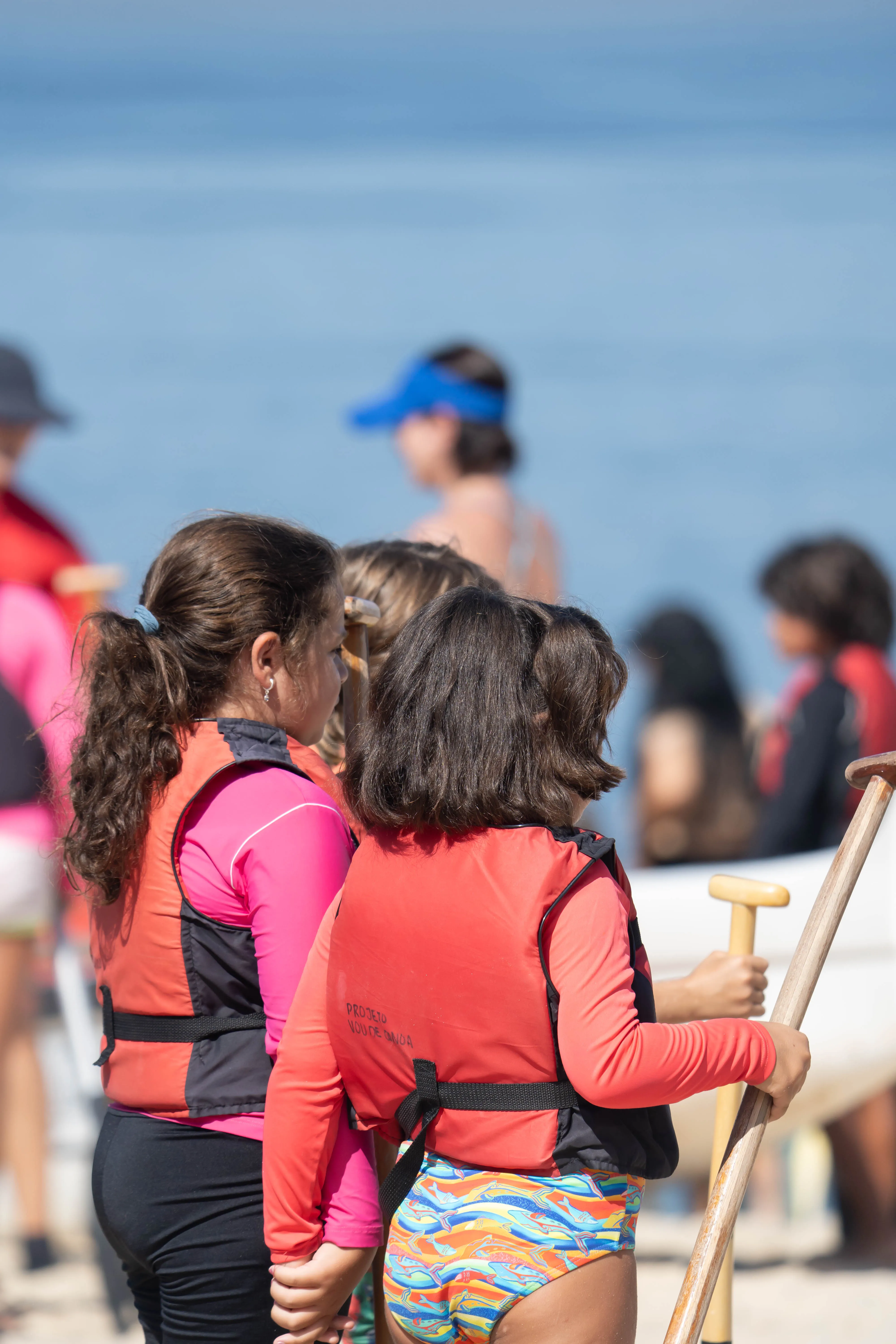 Crianças de costas, com coletes, olhando o mar aberto ao lado da canoa polinésia.