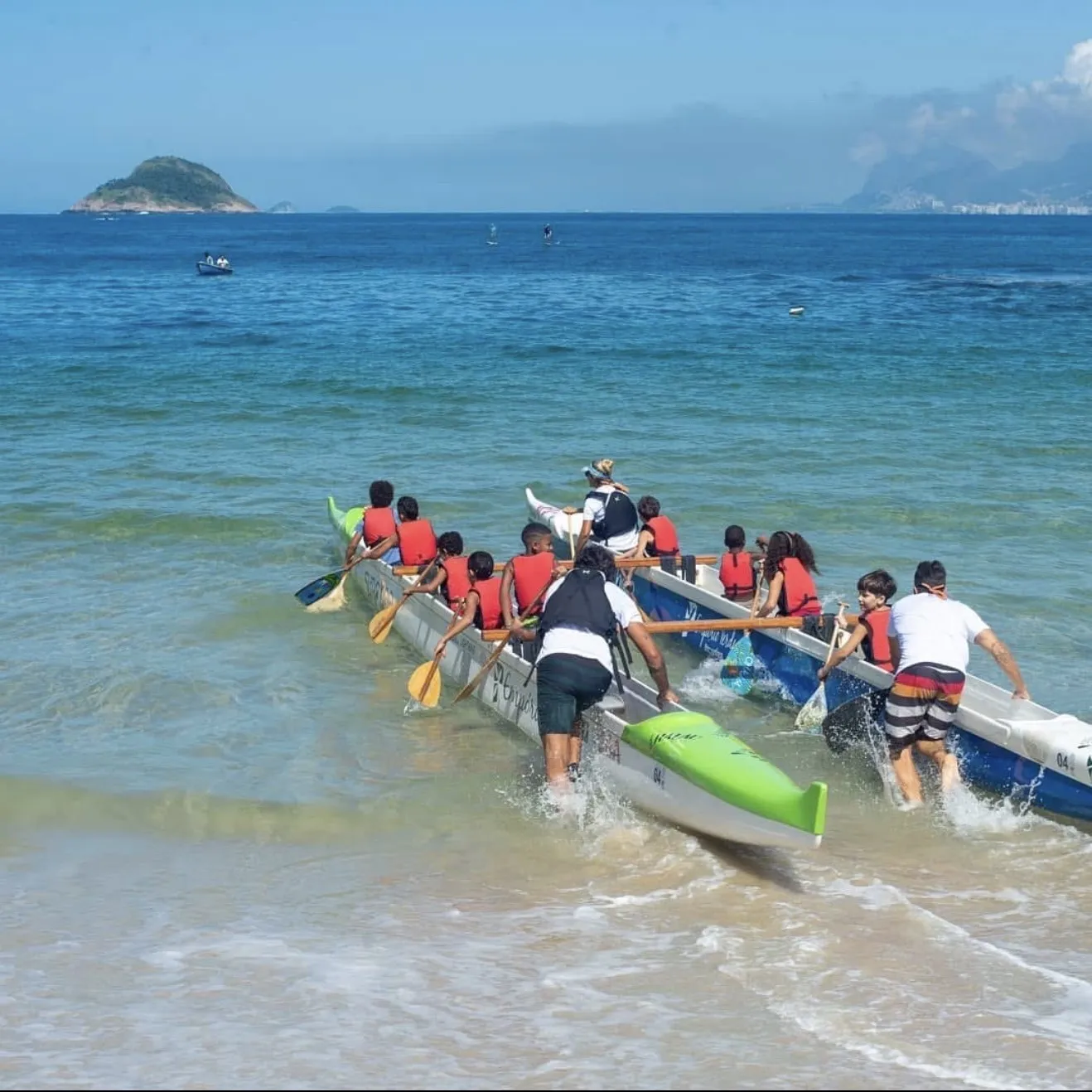 Tripulação de crianças e facilitadores empurra a canoa polinésia para dentro do mar aberto, com ilha e horizonte ao fundo.