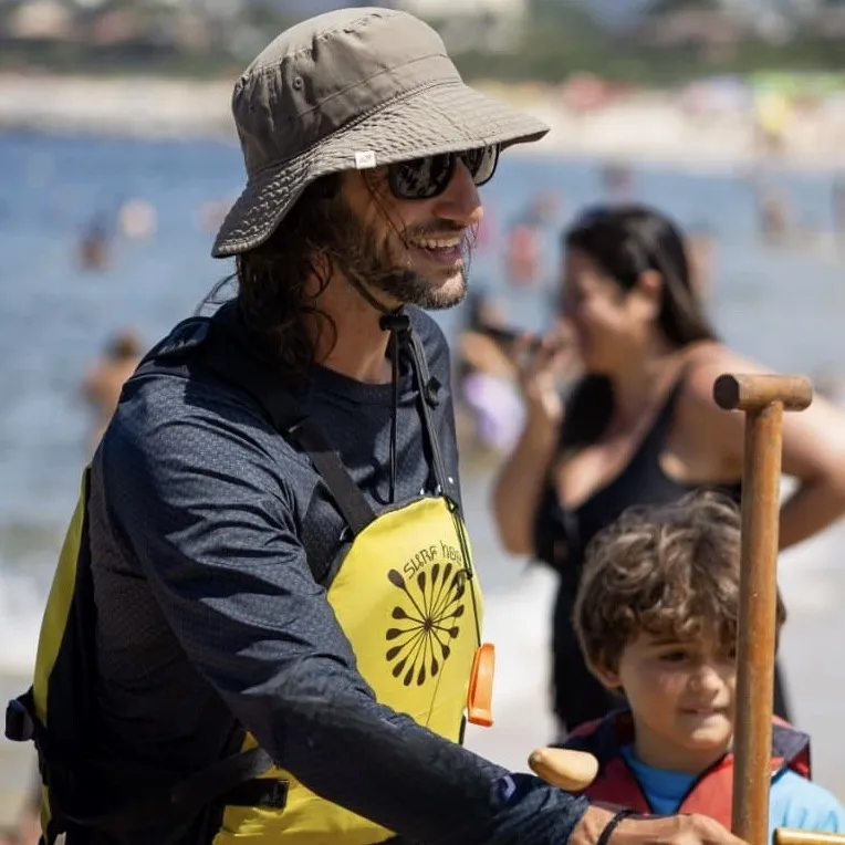 Leonardo Pirozzolo, fundador da Canoa DeVerGente, de chapéu e colete salva-vidas na praia, conduzindo uma criança durante a sessão.