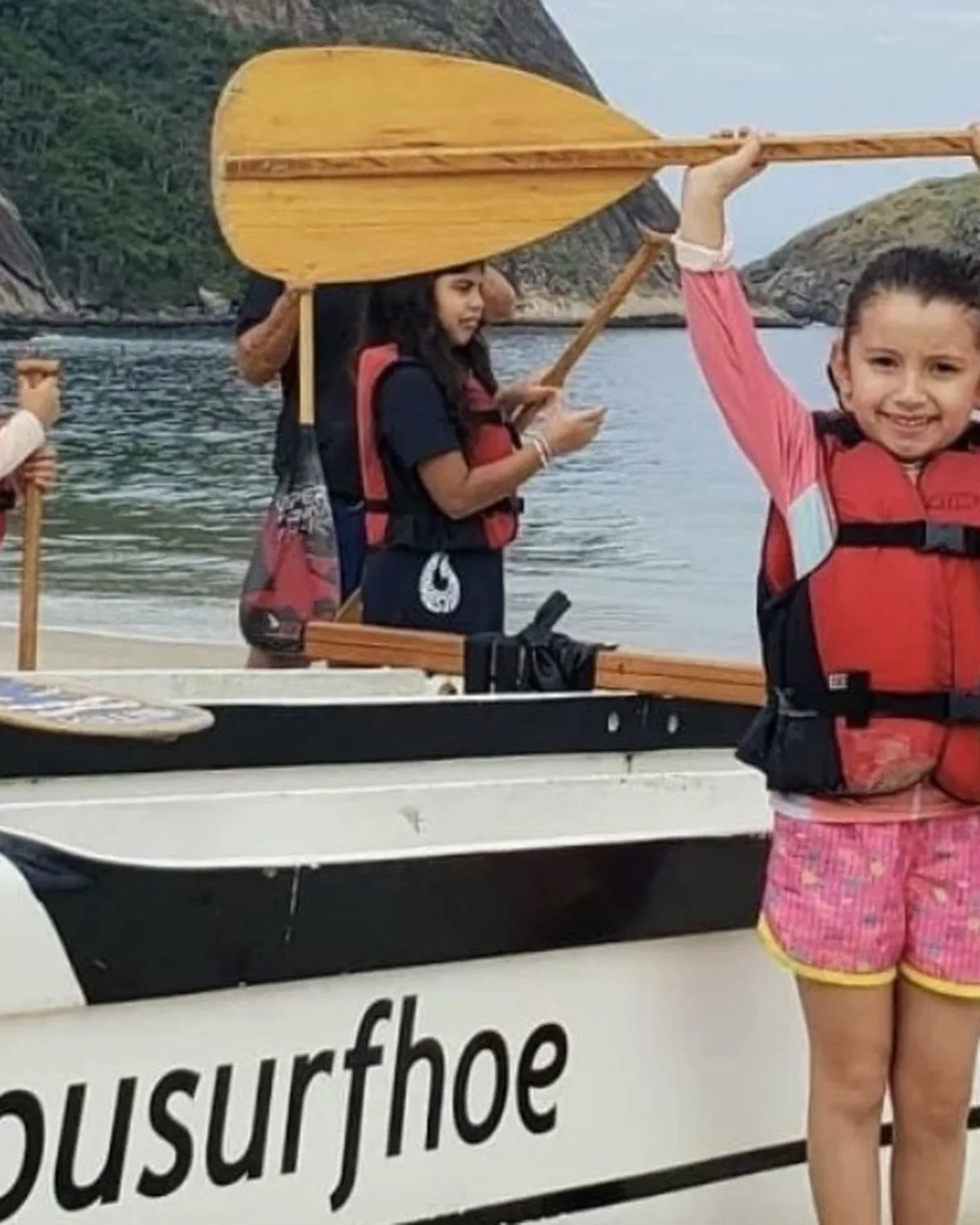 Participante ergue o remo acima da cabeça na praia, ao lado da canoa polinésia.