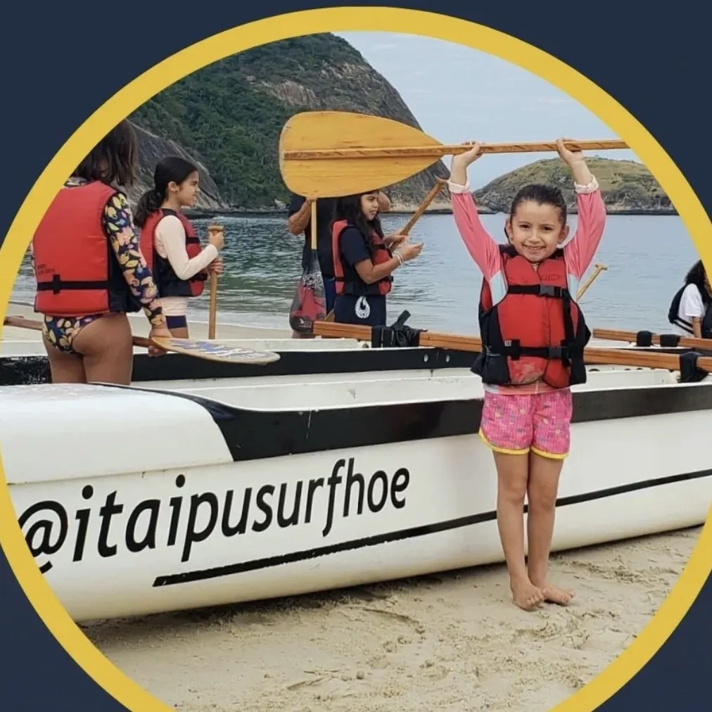Menina sorrindo ergue o remo acima da cabeça na praia, ao lado da canoa polinésia e do restante da turma.