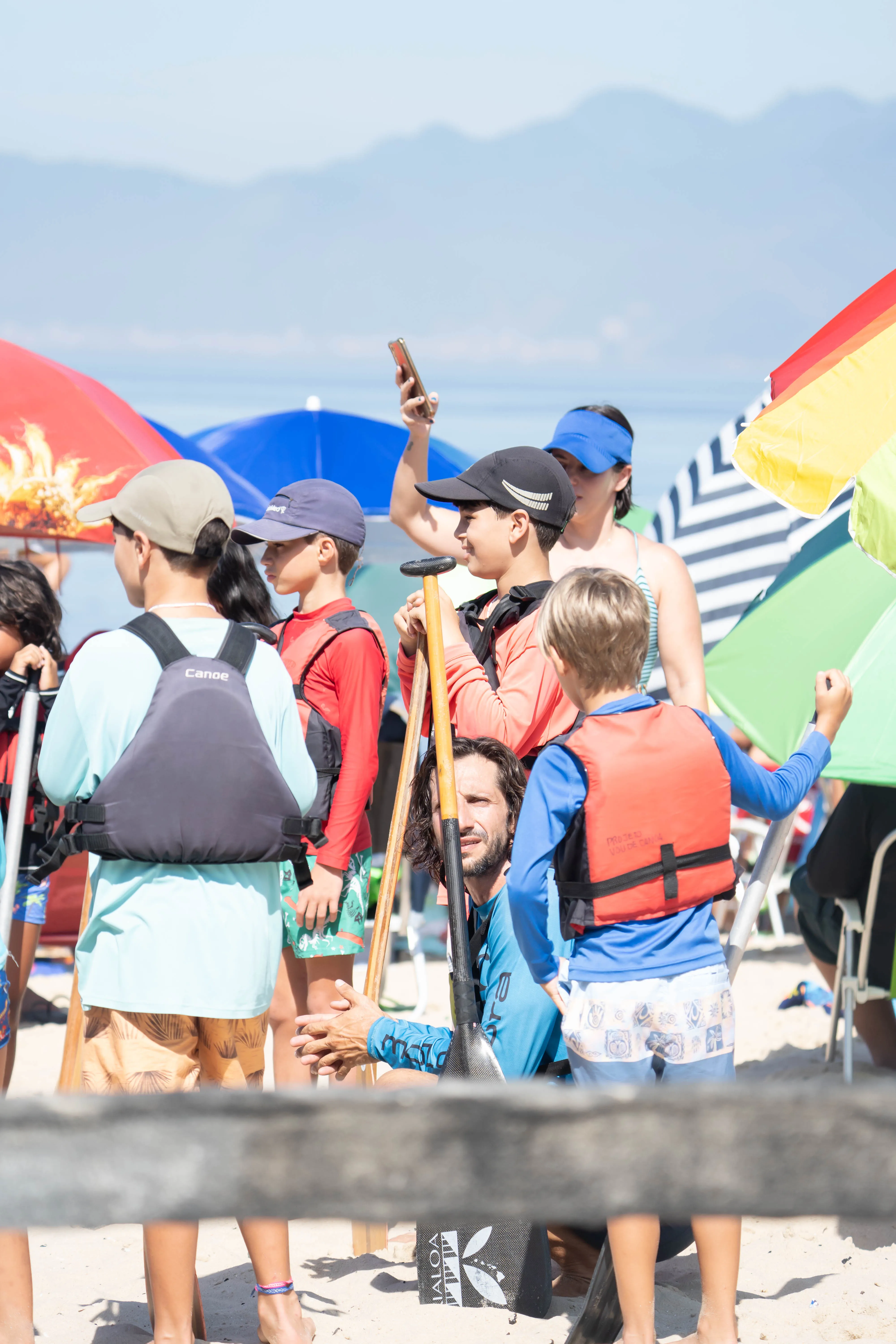 Grupo de crianças e adolescentes de colete salva-vidas, com remos na mão, reunido na areia antes de entrar no mar.