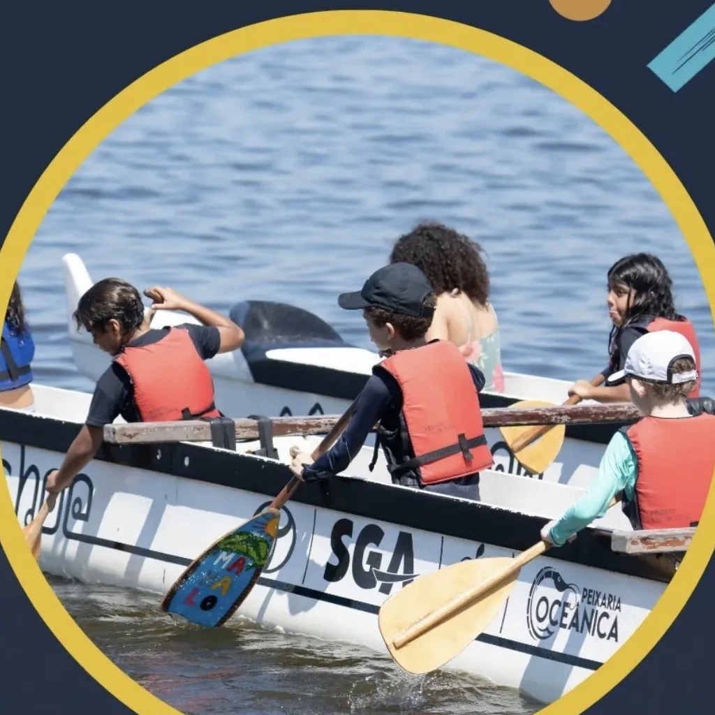 Close de crianças remando na canoa polinésia, com os remos entrando na água no mesmo ritmo.
