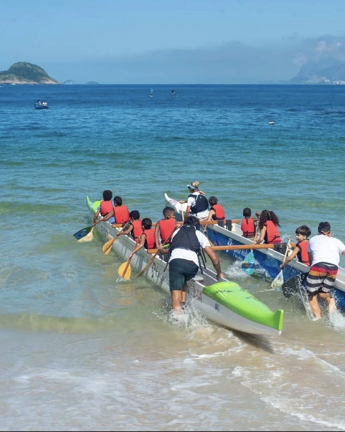Tripulação de crianças e facilitadores empurra a canoa polinésia para dentro do mar aberto, com ilha e horizonte ao fundo.