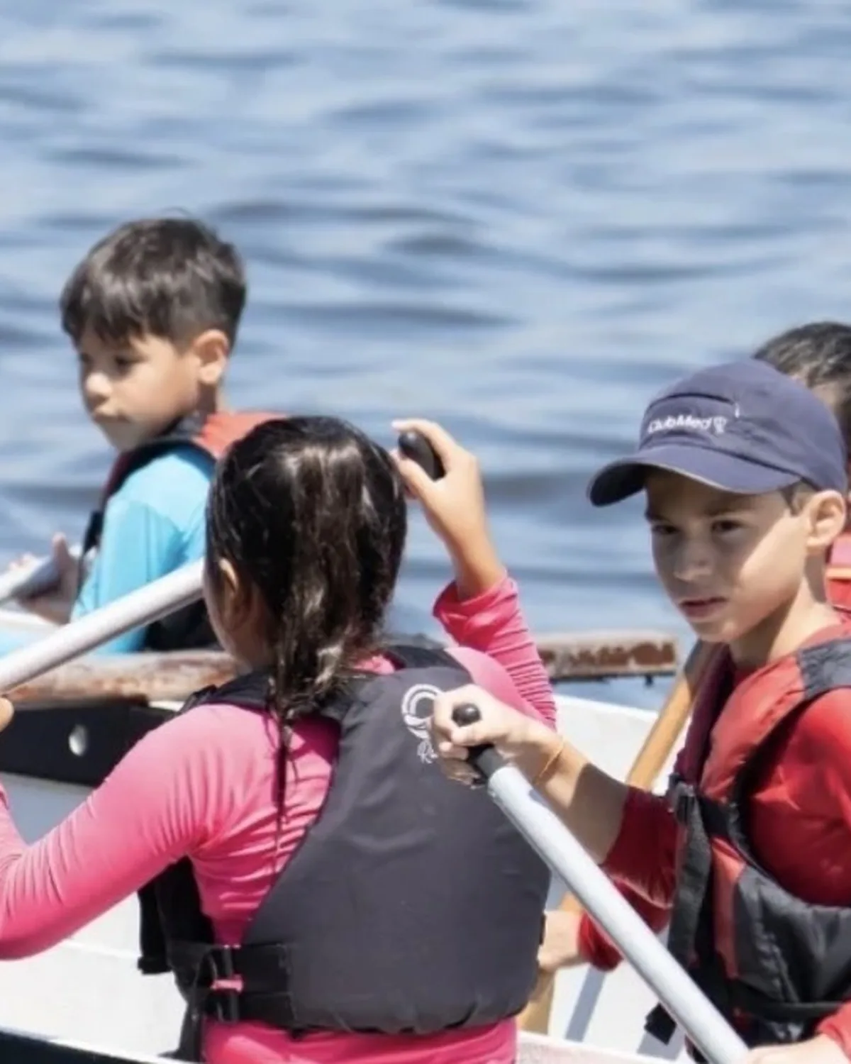 Crianças de colete salva-vidas remando lado a lado dentro da canoa polinésia, cada uma olhando para uma direção diferente.