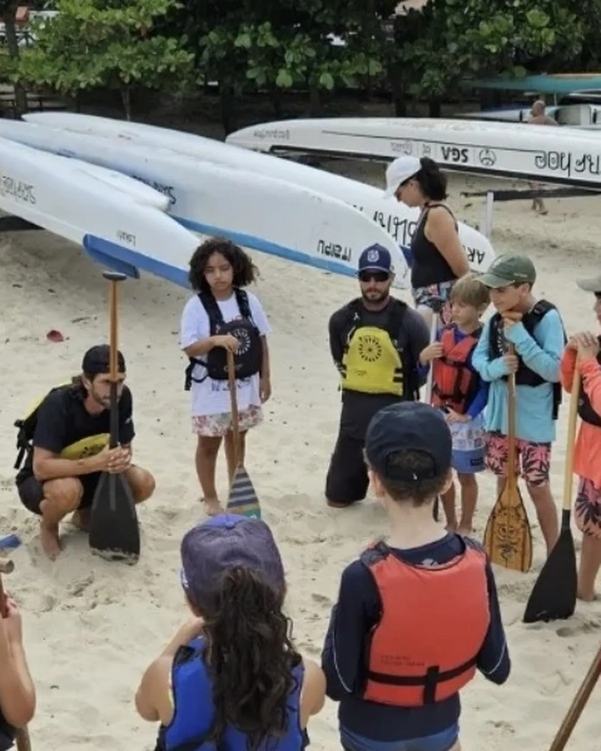 Facilitador agachado na areia conversa com uma roda de crianças de colete salva-vidas e remo na mão, antes de entrar no mar.